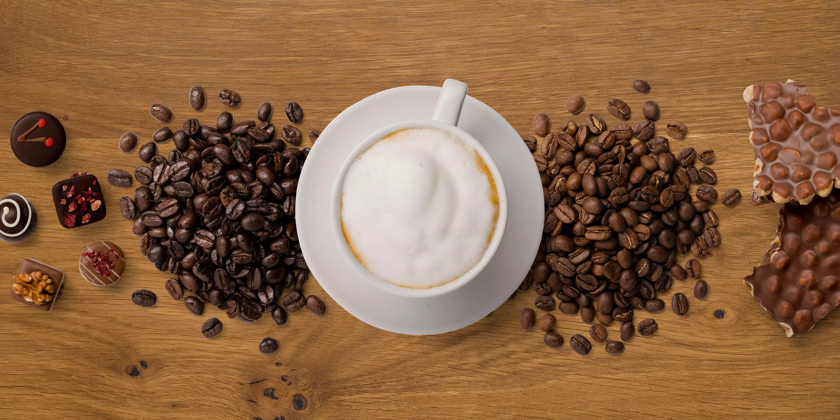 Von oben fotografierte Cappuccino-Tasse, flankiert von Kaffeebohnen, Pralinen und Nuss-Schokolade