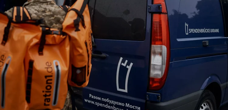 Hilfsgüter in orangefarbenen Taschen vor einem dunkelblauen Transporter mit dem Aufdruck 'Spendenbrücke Ukraine'.