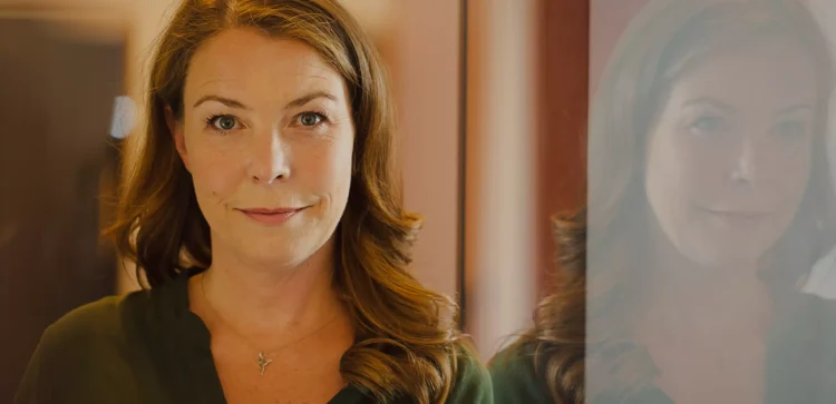 Leane Zaborowski, lächelnd für ein Porträt mit ihrer Spiegelung in einer Glasscheibe, in einem olivgrünen Top.