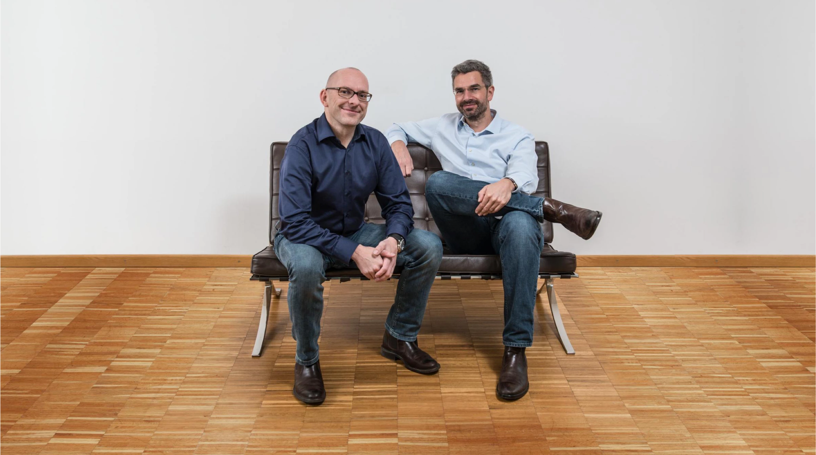 Die Gründer von SOFA53NEUN, Carlo Krüger und Niels Bünemann, sitzend auf einem Designer-Sofa im minimalistischen Stil in einem Raum mit Holzboden.